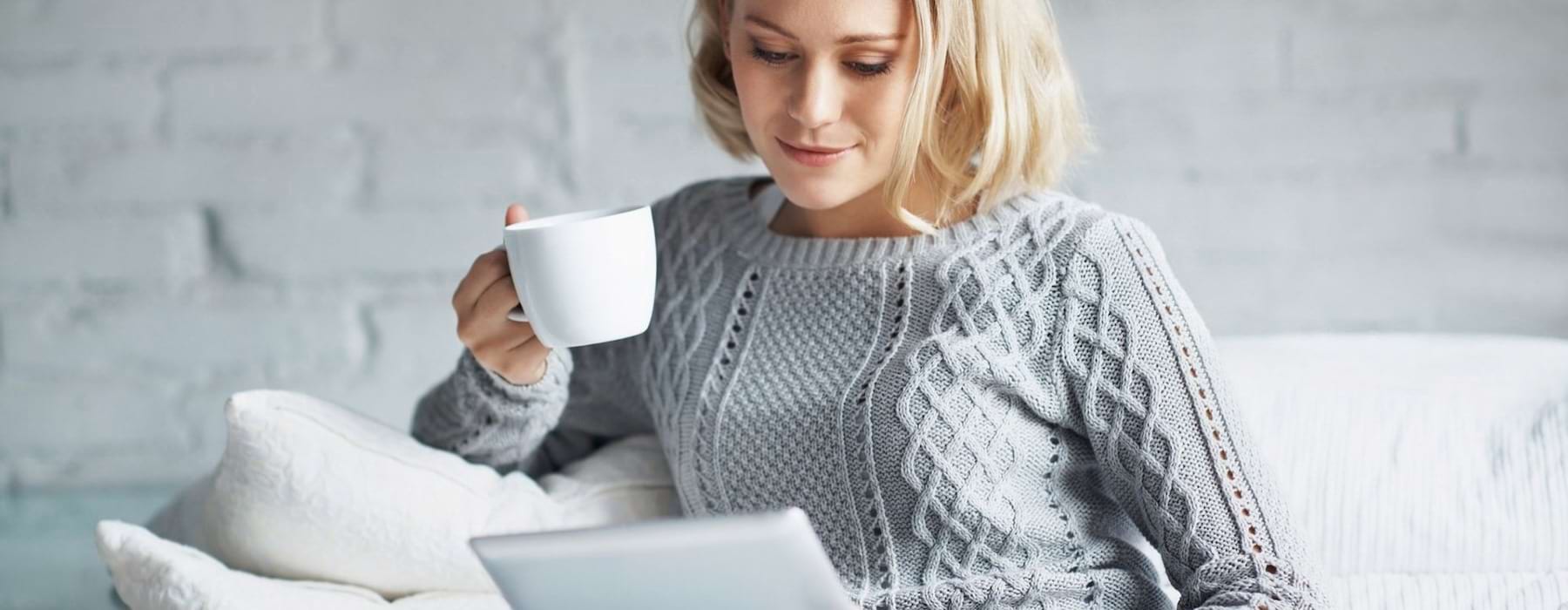 a woman sits on a couch with a cup of coffee and looks at her tablet