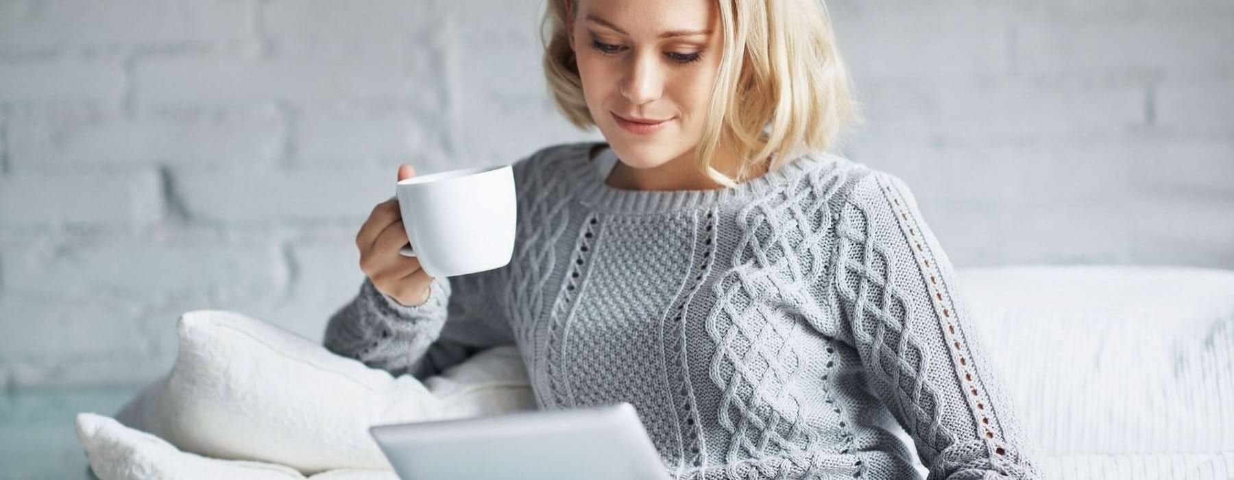 a woman sits on a couch with a cup of coffee and looks at her tablet